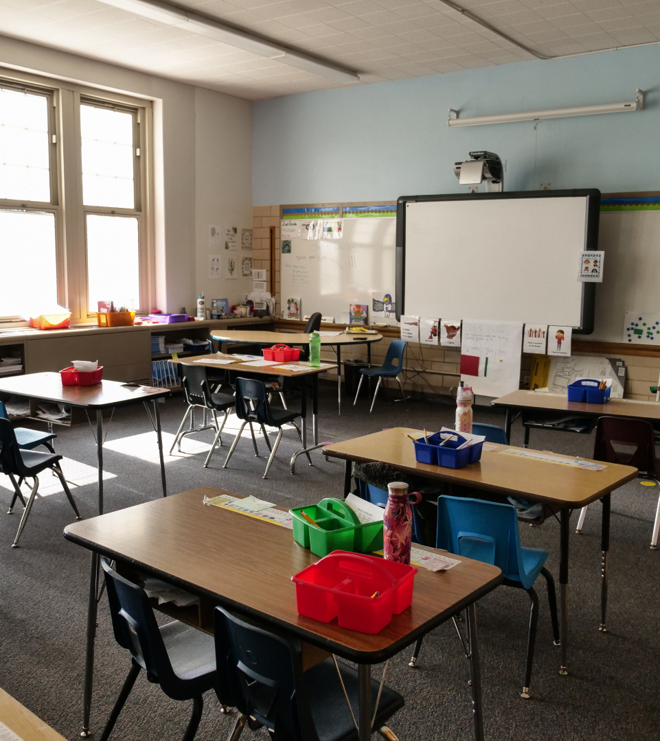 An empty classroom with student desks, various school supplies on the desk, and lights dimmed